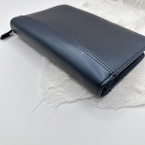 May include: A black, rectangular zippered wallet made of a smooth material. The wallet has a stitched seam down the center and a black zipper pull. The wallet is resting on a white, textured surface.
