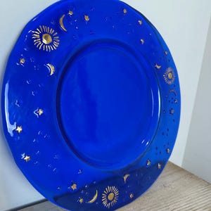 May include: A circular cobalt blue glass plate featuring a raised gold-coloured celestial pattern. The design includes multiple suns, crescent moons, and various sizes of stars scattered across the rim and surface of the vibrant blue glass.