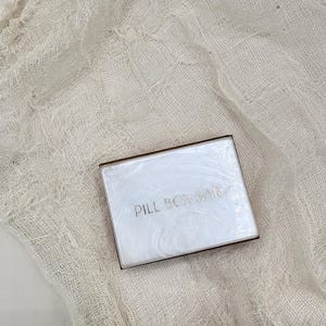 May include: A rectangular pill box with a pearlescent white surface and the text "PILL BOX BAR" in silver lettering. The box has a thin, brown border and rests on a cream-colored, textured fabric.