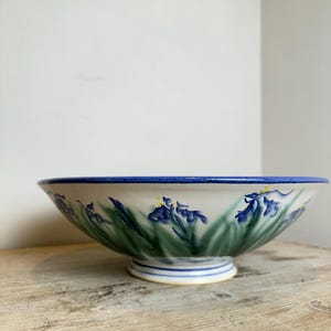 May include: A ceramic bowl with a white base featuring a hand-painted pattern of blue iris flowers and green leaves around the exterior. The bowl has a wide, flared rim with a solid blue edge and a raised circular foot base with blue line details.