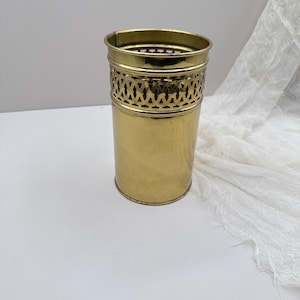 May include: A cylindrical brass container with a hammered texture and a decorative openwork band. The top and bottom rims are smooth, and the container stands on a white surface. The container is likely used for storage or display.