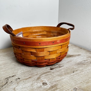 Può includere: Un cesto rotondo in legno intrecciato con una finitura naturale marrone chiaro e una fascia orizzontale rossa a contrasto vicino al bordo superiore. Il cesto include due manici in pelle marrone scuro fissati ai lati e piccoli rivetti.