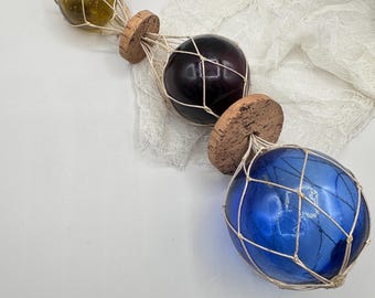 May include: Three glass fishing floats in shades of green, purple, and blue, each encased in a white net and attached to a cork base. The floats are arranged diagonally against a white background, evoking a nautical theme.