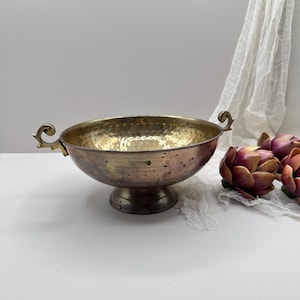 Puede incluir: Un cuenco de metal martillado con base de pedestal y asas decorativas. El cuenco tiene un interior dorado y un exterior de bronce. Las asas son ornamentadas y curvadas. El cuenco es probablemente para fines decorativos.