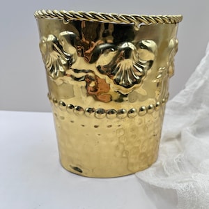 Könnte beinhalten: Ein goldfarbener Metalleimer mit gehämmertem Muster und einem seilartigen Rand. Der Eimer hat geprägte Muscheldekore und eine Reihe kleiner, runder Verzierungen. Das Gesamtdesign ist verziert und dekorativ.