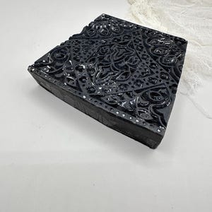 May include: A black, rectangular block featuring a detailed, raised floral design. The edges are slightly worn, and the block has a matte finish. Small silver dots are along the edges. It appears to be a decorative item or a stamp.