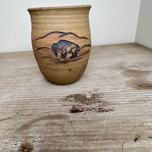 Puede incluir: Un jarrón de cerámica de color marrón claro con una superficie texturizada. El jarrón presenta un diseño tallado de un bisonte en un paisaje. El bisonte está coloreado en tonos azules y marrones. El jarrón tiene un borde ligeramente ensanchado.