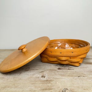 Può includere: Un cestino rotondo in legno intrecciato color marrone chiaro con un coperchio liscio dotato di pomello centrale. Il cestino presenta un rivestimento interno in plastica trasparente ed è realizzato con strisce di legno sovrapposte e chiodini.