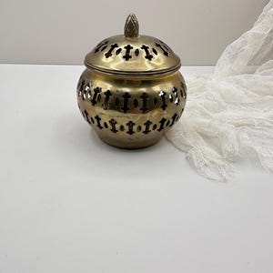 May include: A vintage brass incense burner with a lid. The round body features decorative cut-out patterns and a textured surface. The lid has similar cut-outs and a small, textured knob. The burner is designed to hold incense.