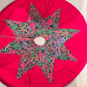May include: A red Christmas tree skirt with a star-shaped design. The star is made of green fabric with a pattern of colorful bows. The skirt has a circular shape with a hole in the center for the tree trunk.
