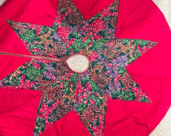 May include: A red Christmas tree skirt with a star-shaped design. The star is made of green fabric with a pattern of colorful bows. The skirt has a circular shape with a hole in the center for the tree trunk.
