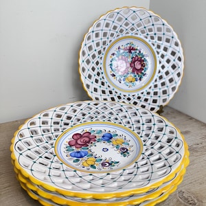 May include: A stack of four white ceramic plates featuring a lattice-style openwork border with a yellow scalloped edge. The center of each plate displays a hand-painted floral motif in shades of red, blue, yellow, and green with thin circular lines.