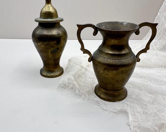 May include: Two antique brass vessels. One is a small lidded urn with a pointed finial. The other is a handled pitcher with a wide opening. Both have a tarnished, aged appearance, suggesting they are vintage or antique decorative items.