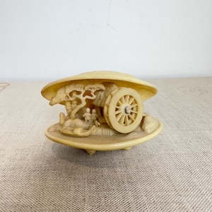 Könnte beinhalten: Eine cremefarbene, muschelförmige Schmuckschatulle mit einer detaillierten, geschnitzten Reliefszene im Inneren. Die Szene zeigt einen kleinen Wagen mit einem großen Speichenrad, einen Baum und zwei kleine Figuren in einem hellen Farbton.
