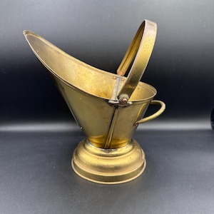 May include: A brass coal scuttle with a handle and a round base. The scuttle is a traditional household item used to carry coal.