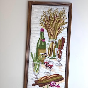 May include: A framed needlepoint wall hanging depicting a still life scene with a green wine bottle, a glass vase filled with wheat stalks, two wine glasses containing red liquid, a glass with green herbs, a loaf of bread, and a red onion.