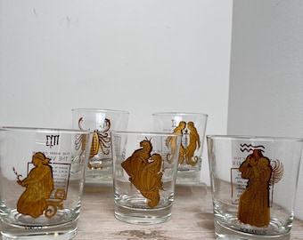May include: A set of five clear glass tumblers featuring gold-colored zodiac illustrations and text. Each glass displays a specific astrological sign, including Virgo, Scorpio, Pisces, Gemini, and Aquarius, with corresponding figures and symbols printed.