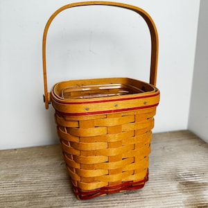 May include: A square woven wood basket with a light brown finish and a single arched handle. The basket features a horizontal red accent stripe near the top rim and a matching red accent stripe along the base, with a clear plastic liner inside.