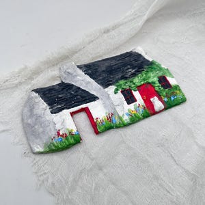 May include: A miniature, hand-painted cottage with a black roof, white walls, and a red door. The cottage is decorated with green foliage, colourful flowers, and a small white cat. The artwork is set against a white, textured backdrop.