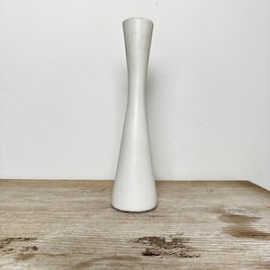 May include: A tall, slender, matte white candlestick holder with a smooth, tapered silhouette. The object features a wide, circular base that narrows into a thin, elongated neck before slightly widening again at the top opening for a candle.