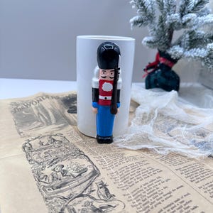 Może przedstawiać: Ceramiczna figurka świątecznego dziadka do orzechów w mundurze żołnierza, z czarnym kapeluszem, czerwoną kurtką i niebieskimi spodniami, stoi przed białym kubkiem. Figurka umieszczona jest na zabytkowej gazecie z ilustracjami i tekstem.