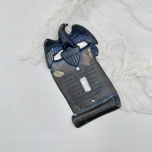 Puede incluir: Una cubierta decorativa para interruptor de luz con un diseño de águila y escudo. La cubierta es de color bronce y presenta una cita de Thomas Jefferson. El águila está posada sobre la abertura del interruptor, con la cita debajo.