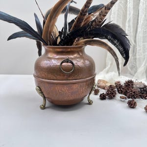 以下が含まれることがあります： 真鍮のリングハンドルと真鍮の足が付いた、ハンマー加工された銅製の鍋型の花瓶。黒、茶色、タンの色合いの羽で満たされています。家の装飾用の装飾品。