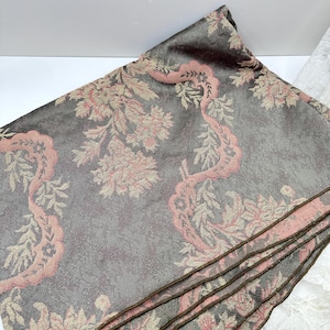 May include: Folded gray fabric with a floral pattern in pink and beige. The fabric has a textured appearance and a dark brown border. The floral design features stylized leaves and flowers. The fabric is folded over, revealing multiple layers.