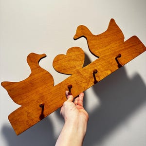 May include: A wooden wall-mounted rack featuring a natural brown finish, shaped with two duck silhouettes and a central heart. The piece includes four small, evenly spaced wooden pegs extending from the front for hanging items.