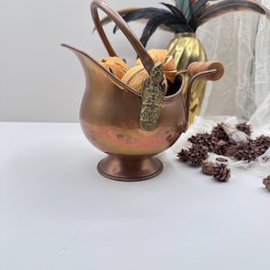 May include: A copper-colored metal pitcher with a handle and a decorative clasp. The pitcher is filled with dried orange slices. A gold vase with feathers and pinecones are in the background. The pitcher is a decorative item.
