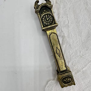 Op de afbeelding: Een messingkleurige flesopener in de vorm van een klok in vintage-stijl. De opener heeft een gedetailleerde wijzerplaat met Romeinse cijfers en sierlijke details. De langwerpige handgreep heeft een ruitvormig accent en een leliebasis. Ongeveer 15 cm lang.
