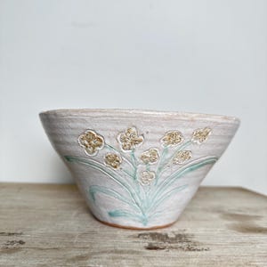 Puede incluir: Un cuenco de cerámica hecho a mano con un acabado mate texturizado en color blanco roto. El exterior presenta un diseño floral en relieve con tallos y hojas de color verde claro, acentuado por pequeñas flores de color tostado.