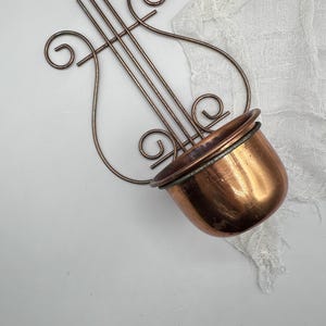May include: A copper-colored, lyre-shaped wall planter with a small, round pot. The planter has a decorative design with curved lines and swirls. The pot is angled and appears to be made of copper.