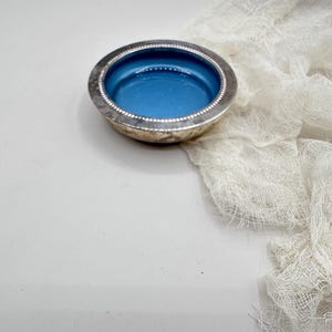 May include: A small, round silver dish with a bright blue interior. The rim is detailed with a beaded design. A white, gauzy fabric is draped in the background, creating a soft contrast to the dish's metallic sheen.
