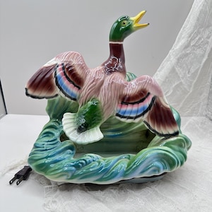May include: A ceramic duck figurine with outstretched wings, featuring a green head, brown body, and colorful wing details. The duck sits atop a wave-like base with a plug attached, suggesting it may be a lamp or decorative piece.