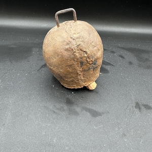 May include: A rustic, brown cowbell with a metal handle. The bell has a worn, aged appearance and a wooden base.