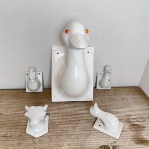 May include: A collection of five white ceramic wall hooks featuring animal motifs. The set includes a large central goose head, two smaller goose figures, a cow head, and a duck figure. Each piece is mounted on a rectangular or shaped base.