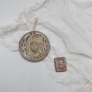 Puede incluir: Dos adornos de cerámica. Uno es redondo con un diseño de pájaro y una combinación de colores beige y marrón. El otro es rectangular con un diseño de barco. Ambos tienen un agujero para colgar y están sobre una tela blanca texturizada.