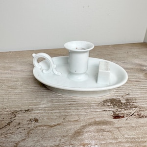 Peut inclure: Un bougeoir en céramique blanche avec une poignée et un petit bloc carré. Le support a une base ronde et une coupelle centrale pour une bougie. La poignée a un design décoratif et incurvé.