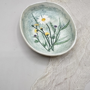 May include: Oval ceramic bowl with a hand-painted floral design. The bowl features a light blue-green background with a white daisy, yellow flowers, and green leaves. The rim is white, and the bowl is approximately 8 inches long.