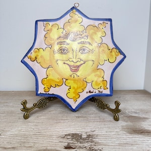 May include: A star-shaped ceramic wall plaque featuring a hand-painted yellow sun with a smiling face against a light pink background. The plaque has a blue border and the text "Made in Italy" is written in black script on the lower right side.