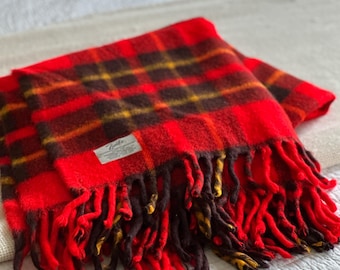 May include: A red and brown plaid scarf with a fringed edge. The scarf has a label in the corner. The plaid pattern includes yellow and black accents. The scarf is folded on a white surface.