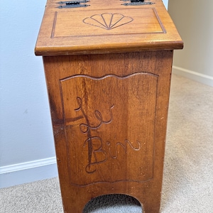 May include: A medium-brown wooden bin with a hinged lid featuring two black metal hinges. The lid has a carved shell design, and the front panel features a carved wavy border and the text "Le Bin" in a cursive script. The base has an arched cutout.