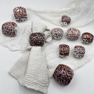 May include: A selection of decorative napkin rings featuring a speckled brown and cream pattern. The napkin rings are displayed on a cream-coloured cloth, with a folded napkin. The design resembles a seashell.