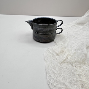 May include: A small, antique silver-toned creamer with a spout and two small handles. The creamer has horizontal banded detailing around the body. The background is a white surface with a white, gauzy fabric draped across it.