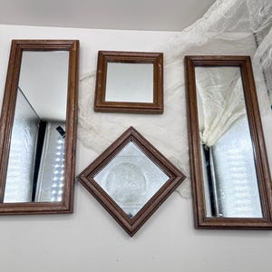 May include: Four decorative mirrors with brown wooden frames. The mirrors are rectangular, square, and diamond-shaped. The mirrors are arranged on a white surface with a white fabric draped across the top.