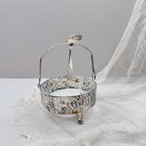 May include: A silver-colored, vintage sugar bowl with a handle and a spoon. The bowl has a decorative, openwork design around the sides and sits on three small feet. The spoon rests in the center of the handle.