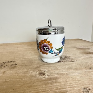 May include: A white ceramic egg cup with a polished silver-tone metal lid featuring a small vertical ring handle. The exterior of the cup is decorated with a colorful floral pattern in shades of orange, purple, blue, and green on a white background.
