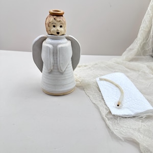May include: A ceramic angel figurine with a white body, wings, and a detailed face. The angel has a brown halo and is standing on a light brown base. A white tag with a string is next to the angel.
