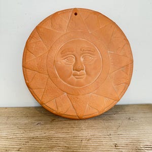 Könnte beinhalten: Eine kreisförmige Wandplatte aus Terrakotta mit einem zentralen Sonnengesicht mit geschlossenen Augen und einem ruhigen Ausdruck. Die Sonne ist von dreieckigen Strahlen umgeben, die mit geprägten Spiralmustern verziert sind. Oben befindet sich ein Loch zum Aufhängen.